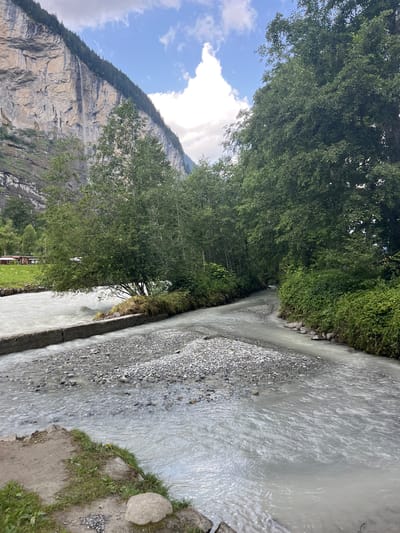 The image depicts a scenic view in Wengen, Switzerland. A wide, milky-white river flows through a valley, bordered by lush green trees and a steep, rocky cliff face. The riverbed is visible in places, with numerous grey and white pebbles. A concrete structure, possibly a weir or a retaining wall, runs along the left bank of the river in the foreground. On the far left bank, a small patch of grass and gravel is visible, with a single large, light-colored rock. In the middle ground, trees line both sides of the river, with a dense forest on the right. The cliff face, covered in scattered trees and vegetation, dominates the background on the left. A thin waterfall cascades down the cliff face. The sky is partly cloudy with patches of blue. There are no people or animals visible in the image.