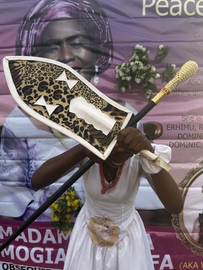 The image presents a close-up view of a person holding ceremonial items against a vibrant banner backdrop. The individual, dressed in a white garment with a sheer overlay and golden embroidery, wears an orange beaded necklace. Their hands are adorned with white ruffled wrist wraps. They firmly grasp a long, dark staff featuring an ornate golden handle intricately embellished with jewels. In the other hand, they hold a shield covered in a distinctive leopard print fabric, accented with white trim and appliqued white bow-tie and cloud-like shapes.

The background is a large purple and pink banner or poster, indicating a special event. Visible text on the banner includes "Peace," "Mother, gra" (likely "grandmother"), "She is," "ERHIMU," "DOMINIC," "MADAM MOGIA," and "OBSEQUIAE," strongly suggesting a memorial service or funeral. A large, sideways portrait of a woman wearing a headwrap is printed on the banner, likely the deceased "Madam Mogia." A part of an ornate gold picture frame is also visible on the banner. Small clusters of artificial flowers, light green/white and yellow, are placed around the scene. The setting appears to be an indoor or sheltered outdoor event in Aghalokpe, Nigeria, during the daytime, filled with cultural and symbolic elements for a solemn commemoration.
