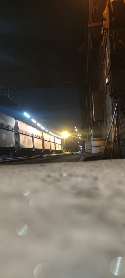 A train of freight cars is parked on railroad tracks at night. The train is illuminated by rows of lights along its side and a bright yellow street lamp further down the track. On the right side of the image, a brick wall is visible with electrical boxes and conduits attached. Two buckets are placed on a surface near the wall. The foreground is blurred with out-of-focus circular bokeh lights, suggesting a low-angle shot possibly from a wet or reflective surface. This scene appears to be in Salzgitter, Germany, likely within an industrial area.