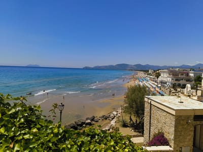 The image shows a panoramic view of the beach and coastline in Sperlonga, Italy. In the foreground, lush green ivy covers a stone wall, with a vintage lamppost visible. Beyond the ivy, a sandy beach stretches along the coastline, dotted with numerous colorful beach umbrellas and loungers. People are scattered across the beach and in the shallow water, enjoying the sunny weather. The turquoise sea gently laps the shore, with small waves breaking. In the distance, a chain of mountains forms the backdrop to the scene, with a hazy island visible on the horizon. White buildings with balconies and rooftops line the edge of the beach, suggesting a developed resort area. The sky is a clear, vibrant blue, indicating a bright and cloudless day.