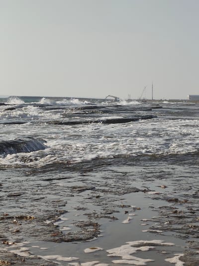 The image depicts a rocky shoreline with waves crashing onto the rocks. In the background, industrial structures are visible, including cranes and a tall tower, suggesting a port or industrial area. The sky is clear and overcast. There are no people or animals visible in the image. The foreground shows a muddy and rocky terrain with some foam from the waves. The scene appears to be located near the city of Saiyn Şapağatov, Kazakhstan, which is known for its industrial port on the Caspian Sea.