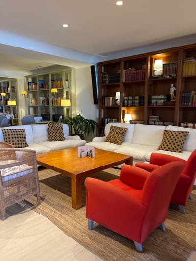 This is an interior photograph of a well-lit lounge area, likely within a hotel or a residential property in Port de Pollença, Spain. The scene is dominated by several pieces of furniture arranged around a central wooden coffee table.

In the foreground, two vibrant red armchairs are positioned on a textured, woven rug. To the left, a wicker armchair with a light-colored cushion is visible. The coffee table, made of light-colored wood with visible grain, is placed centrally on the rug and has a few small items on its surface, possibly menus or decorative objects.

Behind the coffee table, a large, white sectional sofa is the prominent seating arrangement. Two decorative pillows with a geometric pattern in shades of brown and black are placed on the sofa.

The background features extensive wooden bookshelves filled with books. The shelves are organized into multiple sections and extend to the ceiling. There are also decorative lamps and a small statue of a human figure on the shelves. To the left of the main bookshelf, a lighter-colored, open shelving unit, also filled with books and decorative items, is visible. A large potted plant adds a touch of greenery to the scene, positioned near the white sofa. The lighting is warm and inviting, suggesting an evening or interior illumination. The floor transitions from the rug to a light-colored tiled surface.