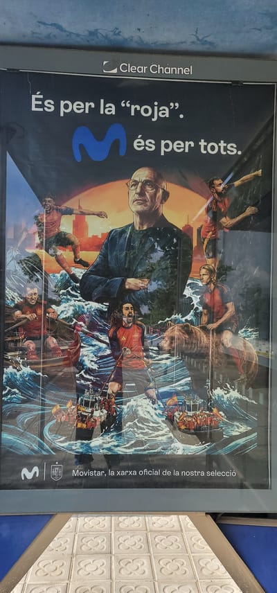 This vertical advertisement poster, seen in Esplugues de Llobregat, Spain, features a vibrant, illustrative depiction of a metaphorical journey or struggle. The central part of the poster shows a dynamic scene where multiple figures, many resembling football players in red and yellow jerseys, are navigating turbulent blue waters. They are engaged in various actions such as rowing boats, pulling ropes, and holding what appear to be flags. A prominent older man with glasses and a serious expression stands in the foreground, seemingly overseeing the activity. Other notable elements include a large brown bear amidst the waves and a cityscape under an orange, sunset or sunrise sky in the background.
The poster contains several textual elements:
- At the top left, the "Clear Channel" logo is visible.
- A prominent slogan in Catalan reads: "És per la 'roja'. I és per tots." which translates to "It's for 'La Roja'. And it's for everyone."
- Towards the bottom right, the Movistar logo is displayed, accompanied by the text "Movistar, la xarxa oficial de la nostra selecció" (Movistar, the official network of our national team).
- The logo of the Spanish Royal Football Federation (RFEF) is also visible near the Movistar branding.
The imagery, particularly the red jerseys and the reference to "La Roja" (a common nickname for the Spanish national football team), strongly links the advertisement to Spanish football. The turbulent waters and determined expressions convey a sense of effort, teamwork, and resilience, likely symbolizing the challenges and collective spirit associated with the national team. The poster's artistic style is heroic and dynamic.