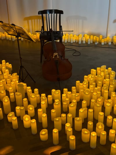 The image depicts a dimly lit indoor scene with a cello and a music stand positioned in front of a dark wooden chair. A large number of flameless LED candles are arranged on the floor, creating a warm, ambient glow. The candles vary in height and some are clustered together, while others are spaced further apart. The background is a plain wall, and the floor appears to be carpeted. The overall atmosphere suggests a concert or performance setting, possibly a memorial service or a special event. The location in Ilyanskiy selskiy Sovet, Belarus is not depicted or suggested by any visual cues within the image.