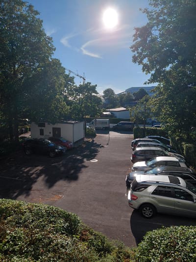 The image depicts an outdoor parking lot on a sunny day. In the foreground, a dense green hedge lines the bottom of the frame. The parking lot itself is paved and contains several vehicles, including a black SUV, a red compact car, and a row of dark-colored cars parked bumper to bumper. A small white building with a dark roof is situated to the left, adjacent to the black SUV and red car. In the background, trees with dense green foliage are visible, casting shadows on the parking lot. Beyond the trees, a modern building with white siding and curved architectural elements can be seen. A construction crane, its jib extended, is visible in the mid-ground to the left, suggesting ongoing construction in the vicinity. The sky is clear blue with a bright sun near the top center, casting lens flare and creating a strong glare. The scene is located in the city of Unterschleißheim, Germany, and appears to be a typical urban or suburban environment on a clear day. There are no people visible in the image.