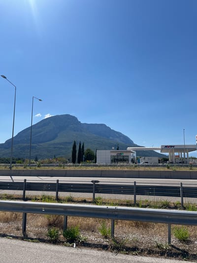 The image depicts a wide, clear blue sky over a mountainous landscape. In the foreground, a highway with a metal guardrail is visible, bordered by dry grass and sparse green vegetation. Beyond the highway, a row of tall, dark green trees stands out against the lighter green hills. Further back, a modern gas station with white structures and prominent signage, including "AVIN" and "Agora," is situated. The gas station appears to be functional with vehicles present. A large, rugged mountain dominates the background, its slopes covered in dense green foliage and rocky outcrops. The overall scene suggests a roadside location in a sunny, rural environment, likely in Greece, given the context of Municipal Unit of Iera Poli Messologhiou. There are no people visible in the frame.