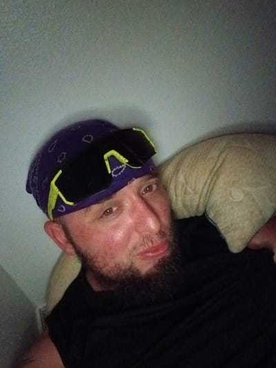The image depicts a man lying on a pillow against a wall. He is wearing a dark bandana and sunglasses with yellow accents. He has a beard and his skin appears flushed. The man is wearing a black shirt. The setting appears to be indoors. The location is unspecified within Clarksville, United States of America.
