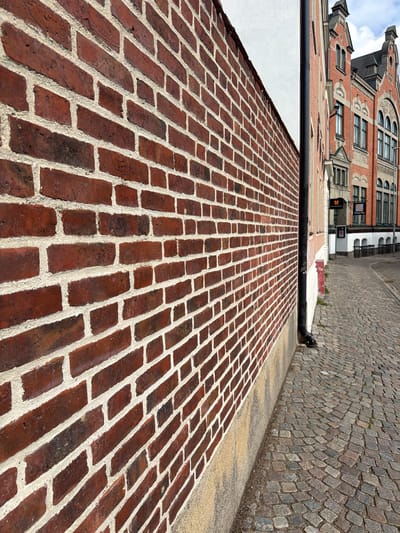 The image shows a cobblestone street in the city of Lund, Sweden, with a prominent red brick wall on the left and historic buildings on the right. The brick wall, made of dark red bricks with light grey mortar, fills a significant portion of the foreground and extends into the background. The street is paved with irregular-shaped grey cobblestones.
In the background, to the right of the brick wall, are several buildings with distinctive architecture. The closest building is an ornate, multi-story structure made of red brick with white trim and dark arched windows. Above it, another building with a dark, sloped roof and pointed architectural features is visible. Several signs are mounted on the buildings, including a dark sign with a crest and another smaller, darker sign. A black downspout runs vertically along the side of the brick wall. The sky is partly cloudy.
No people are visible in the image. The scene is a street-level view, likely taken during the daytime given the lighting. The perspective is from low down, looking diagonally along the street and wall.