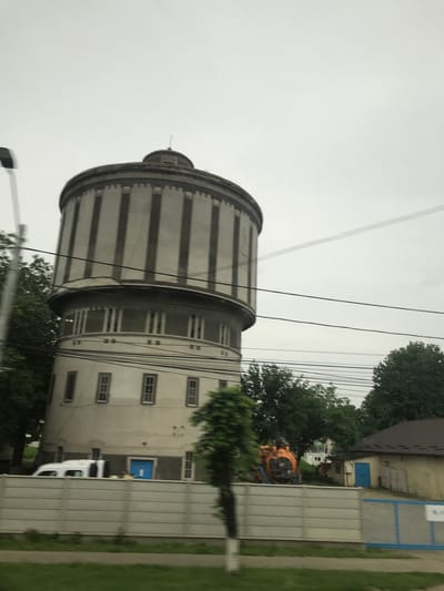 This image captures a multi-tiered cylindrical building, identified as a historic water tower, situated in the city of Roman, Romania. The upper section of the tower features a dark grey cylindrical tank adorned with prominent, regularly spaced vertical dark grey pilasters or decorative bands, capped by a shallow domed roof and a small, slender finial at its apex. Beneath this, a narrower, light grey cylindrical level displays a series of evenly spaced tall, narrow rectangular windows. The widest base section of the tower is light beige, presenting several grey-framed rectangular windows and a recessed blue door at its base on the left side.
In the immediate foreground, a strip of green grass runs along the bottom, bordered by a light grey concrete panel fence that stretches across the scene. A utility pole is partially visible on the far left. Behind the fence, a white panel van is parked towards the left, while further right, a large orange piece of heavy machinery, possibly an excavator or industrial pump, stands next to a smaller, single-story building with a brown-tiled roof and a bright blue door. Abundant green trees are visible around and behind the structures. Numerous black power lines crisscross the middle ground against an overcast, light grey sky. There are no individuals present, and no actions or interactions are depicted.