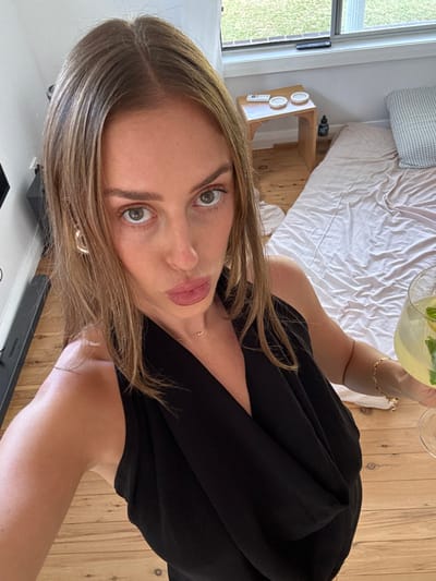 A woman poses for a selfie, holding a drink. She has light brown hair, green eyes, and light skin. She's wearing a black dress and a necklace. A hexagonal earring is visible on her left ear. In the background, there's a bed with white linens, a wooden bedside table, and a window. A wooden floor is beneath her.