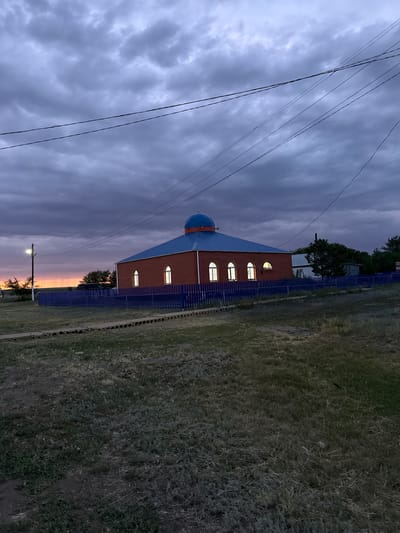 The image depicts a mosque in the city of Өндіріс, Kazakhstan, at dusk. The mosque, constructed of red brick with a blue roof and a blue dome, is illuminated from within, casting a warm glow through its arched windows. A vibrant blue fence encloses the mosque and the surrounding area, which consists of dry grass and sparse vegetation. Power lines crisscross the dramatic, cloudy sky, which transitions from deep purple at the top to hues of orange and pink near the horizon, indicative of a sunset. The overall scene is serene, highlighting the mosque as a focal point against the natural backdrop.
