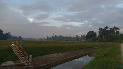 The image depicts a rural scene near Blangtingkeum, Indonesia. A concrete-lined irrigation canal runs through the foreground, with the water reflecting the sky. On the left side of the canal is a yellow metal staircase leading up to a water control structure with pipes and valves. Beyond the canal, there's a field of green and brown vegetation. In the distance, trees line the horizon, blending with the cloudy sky. The light suggests either dawn or dusk. No people are visible. A dirt road is located on the right side of the canal.