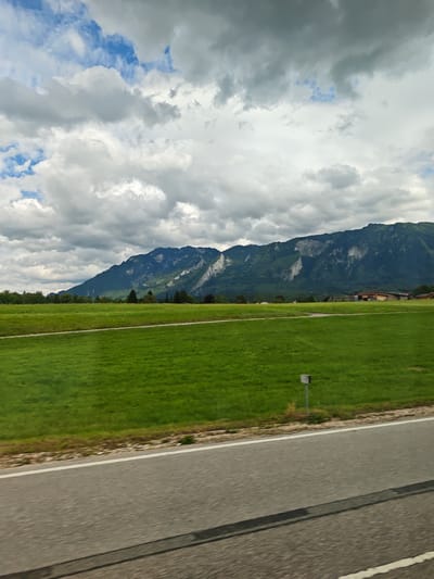 This is a wide-angle, eye-level shot of a pastoral landscape with a cloudy sky over mountains. The foreground features a gray asphalt road with a white line marking its edge and a blurred shadow across it. To the left of the road is a vast, vibrant green field. A thin white line runs across the field, possibly indicating a path or boundary. In the distance, a line of trees forms a dark green border between the field and the mountains. The mountains themselves are steep and imposing, with patches of exposed rock and lush green vegetation. The sky above is filled with dramatic, heavy clouds, with occasional glimpses of blue sky peeking through. The scene evokes a sense of natural beauty and tranquility. Based on the landscape and the architectural style of the buildings in the distance, the location is likely in or around Bad Reichenhall, Germany, known for its alpine scenery.