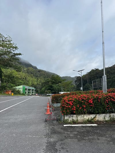 An overcast day in Tambon Wang Prachan, Thailand, presents a view of a paved parking lot with a white lane marking. To the right, a vibrant hedge of red ixora flowers borders a concrete planter. Beyond the flowers, a tall metal pole with a solar-powered street lamp stands, accompanied by a wire mesh fence. In the background, a lush green forest covers a misty mountain slope. A low-rise, green building with a grey roof is visible to the left of the parking lot, next to a small wooden structure and a yellow utility box. A white truck is parked in the distance. Orange traffic cones and a metal frame with wheels are situated near the edge of the parking lot.