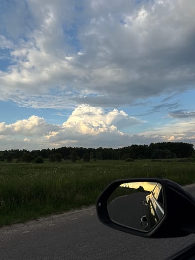 The image captures a serene landscape from the perspective of a moving vehicle, likely during sunset or sunrise. The scene features a dynamic sky, a green field, a line of trees, and a road, with a prominent car side mirror in the foreground.

The sky is a central element, displaying a mix of light blue patches and varied cloud formations. Bright white, voluminous cumulus clouds are illuminated by direct sunlight, while other areas are covered by darker, grey clouds. Distinct crepuscular rays are visible, fanning out from behind a large cloud formation, indicative of the sun being low on the horizon.

Below the sky, a verdant green grassy field stretches across the midground, dotted with small, white wildflowers. A dense, dark green line of trees forms a clear boundary between the field and the distant sky, suggesting a rural or countryside setting.

To the right, an asphalt road runs alongside the grassy verge. In the lower right corner, a black car side mirror is visible, providing a reflection of the scene behind the vehicle. The mirror vividly reflects a bright, golden light, strongly suggesting the sun's position at either dawn or dusk, casting a warm glow over the receding road and the vehicle's bodywork.

No people or visible text are present in the image. The overall impression is one of peaceful travel through a natural landscape, consistent with the given location context of Nadezhda, Russia.