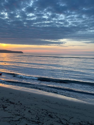 The image depicts a beach scene at sunset. The sky is filled with dramatic clouds, tinged with hues of orange and pink, suggesting the sun is low on the horizon. The ocean is calm, with gentle waves lapping at the shore. The wet sand of the beach reflects the warm light of the sunset, creating a shimmering effect. Scattered seaweed and debris are visible on the darker, drier sand further up the beach. In the distance, a silhouette of land or a hill can be seen against the vibrant sky. The overall mood is serene and peaceful, characteristic of a sunset on a beach. The image is oriented vertically, as if taken with a phone held in portrait mode.