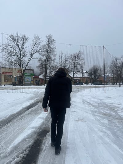 The image depicts an outdoor, snow-covered urban environment in Staryi Saltiv, Ukraine, under an overcast sky. A person, viewed from behind, is the primary subject in the foreground, walking away from the camera toward the center of the frame. This individual is dressed in a black hooded puffer jacket, dark trousers, and dark footwear, and holds a light-colored, rectangular object in their left hand.
The ground is covered in packed snow and ice, exhibiting clear tire tracks and footprints. Along the left side of the midground, several commercial structures are visible. A white building displays signs in Ukrainian, prominently featuring "САЛТІВСЬКИЙ М'ЯСОКОМБІНАТ" (Saltiv Meat Processing Plant) and "АПТЕКА ОПТИКА + ..." (Pharmacy Optics + ...). A few indistinct figures are present near these buildings. Numerous bare deciduous trees are scattered across the scene.
A long, horizontal string of unlit decorative lights or wires is suspended across the street in the midground, attached to vertical poles. In the background, further to the right, are additional residential-style buildings with visible snow on their roofs, and at least two parked vehicles are identifiable on the snow-covered ground. The scene conveys a cold, winter setting.