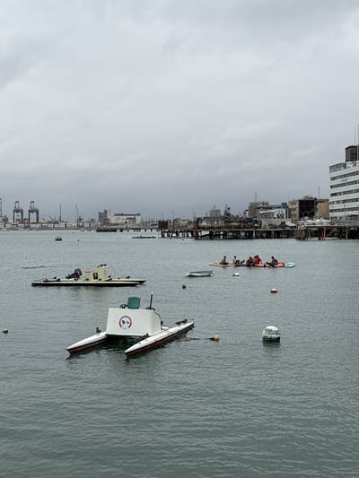 The image depicts a cloudy day in the harbor of La Punta, Peru. In the foreground, a white and red catamaran-style boat with two hulls is moored. Several seagulls are perched on its deck and sides. Further out in the water, a group of people are socializing on a collection of inflatable rafts and paddleboards. A small, white boat with an outboard motor is visible to the left of the catamaran, also with seagulls on board. In the background, a dense urban landscape is visible, characterized by industrial structures, cranes, and large buildings, including a prominent white building with "IMARPE" signage. The water is calm, with a few buoys scattered throughout the harbor.