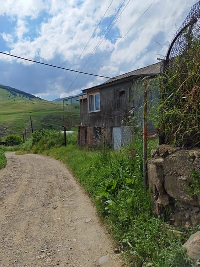 This image depicts a rustic, rural scene in Bazum, Armenia, under a partly cloudy sky. The foreground is dominated by a dirt road winding its way uphill, bordered by lush green grass and vegetation. To the right, a weathered, wooden house with a corrugated roof stands alongside the road. The house has a small balcony with a railing and a large window on the upper floor. Adjacent to the house is a fence made of wire mesh, partially obscured by overgrown plants. A rusty pipe stands vertically near the house, suggesting old infrastructure. In the background, rolling green hills with scattered trees stretch towards a mountainous horizon. The overall atmosphere is serene and suggests a quiet, remote location. The lighting indicates it is daytime, likely afternoon, with the sun intermittently breaking through the clouds. There is no visible text in the image.