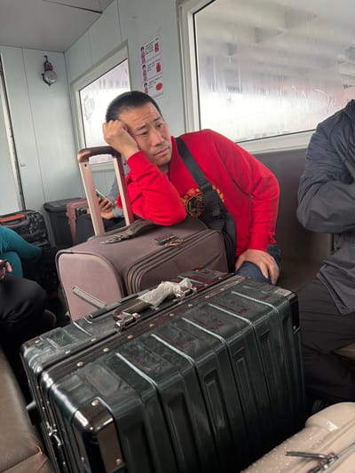 The image captures the interior of a public transport vehicle, likely a train or bus, during a journey. In the center-left, a man of East Asian descent, wearing a red hooded sweatshirt and blue jeans, is seated and resting his head on his hand, looking directly at the camera with a seemingly tired or pensive expression. He has a black shoulder bag slung across his body.

Numerous pieces of luggage are visible, indicating travel. A prominent dark green hard-shell suitcase is in the foreground to the right, with a brown soft-shell suitcase stacked beneath it, positioned next to the man. Other passengers are partially visible, with legs in dark clothing seen in the bottom left and background.

The window on the left side of the frame is obscured by fog or frost, suggesting cold external weather conditions and limited visibility. The vehicle's interior features light-colored walls. Above the window, a white sign with red text and icons is mounted, displaying instructions or warnings. Visible Chinese characters on the sign include "禁止" (forbidden) and "吸烟" (smoking), alongside a crossed-out cigarette icon, indicating a "NO SMOKING" policy. Below the window, a small red and silver bell-like device is affixed to the wall.

Given the location context of Nizhneleninskoe, Russia, which shares a border with China, and the presence of Chinese text on the sign, the scene likely depicts a cross-border journey between Russia and China or travel within a region connected to Chinese culture. The overall mood suggests the routine and weariness of long-distance travel.