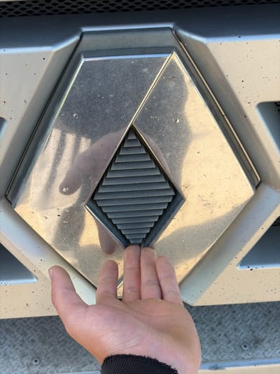 A person's hand is shown reaching towards the grille of a vehicle. The vehicle's grille features a prominent, metallic rhombus-shaped emblem. Inside the emblem is a smaller, ribbed rhombus. The background appears to be outdoors, possibly in Zarech'e, Russia, with a grey, textured surface visible below the vehicle. The lighting suggests it is daytime.