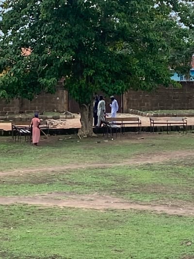The image shows an outdoor scene in Sabo, Nigeria, with a grassy area on the right and a row of benches adjacent to a brick building on the left. Several individuals are seated on the benches. One person is wearing a white garment and appears to be bowing or praying. Another individual is wearing a light green top and dark pants. Farther down the row of benches, another person is wearing a pink dress and a dark-colored head covering. A tree with dense foliage is present to the left of the benches. The ground cover is predominantly grass with visible dirt paths.