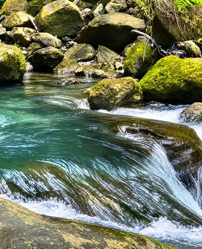 The image depicts a shallow, fast-flowing stream in a rocky, natural environment. The water is a vibrant turquoise color, with white foam indicating movement over submerged rocks and a slight cascade. The stream bed appears to be composed of smooth, rounded stones, some of which are covered in vibrant green moss. The banks of the stream are lined with larger, weathered rocks, also exhibiting mossy growth. In the background, the terrain is densely packed with a variety of rocks, some of which are quite large and angular. Lush green vegetation, including ferns and other leafy plants, can be seen growing among the rocks and on the higher ground, suggesting a humid, shaded environment. The overall impression is of a secluded, untouched natural landscape, likely within a forest or mountainous region. The specific location in the city of Gunung Ambat, Indonesia, is not visually evident from the image itself, as there are no man-made structures or distinct geographical markers that would confirm this locality. The scene is devoid of human presence or any discernible action, focusing solely on the raw elements of nature: water, rocks, and vegetation.