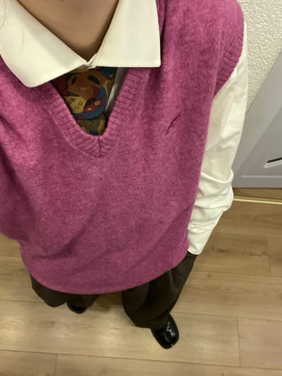 A person is shown wearing brown pants, shiny black shoes, a white collared shirt, a patterned top, and a pink v-neck sweater. The floor is a light brown, and a white cabinet is visible in the upper left. The photo is taken from above, focusing on the person's upper body and legs.
