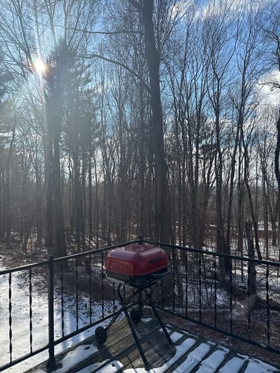 This image depicts a serene outdoor winter scene, likely a backyard or deck area in Charlton, United States. The setting is dominated by a dense forest of bare deciduous trees in the background, their intricate branches creating a delicate pattern against a clear, bright blue sky with sparse white clouds.
In the mid-ground, a black metal railing separates the immediate foreground from the wooded expanse. Positioned behind this railing, towards the right side of the frame, is a prominent red, kettle-style grill with wheels. The ground surface, visible on the right, appears to be a wooden deck or patio, partially covered in a layer of pristine white snow, confirming the winter season.
The sun is shining brightly from the lower left portion of the image, casting a strong glare and lens flare as its rays filter through the skeletal tree branches. This suggests a sunny day, likely late morning or early afternoon. No people or specific activities are present, lending a quiet and still atmosphere to the scene. There is no visible text in the image.