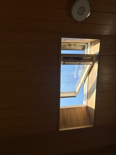 The scene depicts the interior of a room in Hagen, Germany, characterized by light-brown, horizontally oriented wooden paneling covering the walls and ceiling. A rectangular, vertically oriented window, likely a skylight, is positioned on the right side of the frame. The lower section of the window is open, angled inward and upward, revealing a clear blue sky beyond. Strong direct sunlight enters through this opening, illuminating the right side of the window recess and casting bright highlights on the adjacent wooden panels. The left portion of the room and parts of the upper wall remain in shadow. A circular, silver-toned object, possibly a light fixture or ventilation cover, is affixed to the upper wall or ceiling in the top-right quadrant. No individuals or discernible personal items are present.