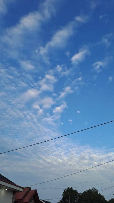 A vertical, medium shot captures a bright blue sky filled with a sparse distribution of fluffy white clouds. A thin, black electrical wire bisects the frame horizontally, running from the left edge towards the right. Below the wire, the upper portions of several rooftops are visible, featuring red tiled roofs and white walls. A cluster of dark green trees with dense foliage occupies the bottom right corner of the image. The scene suggests an outdoor, possibly residential setting, in Lhokawe Teungoh, Indonesia, under a clear sky with scattered clouds. There are no people or animals visible in the frame.