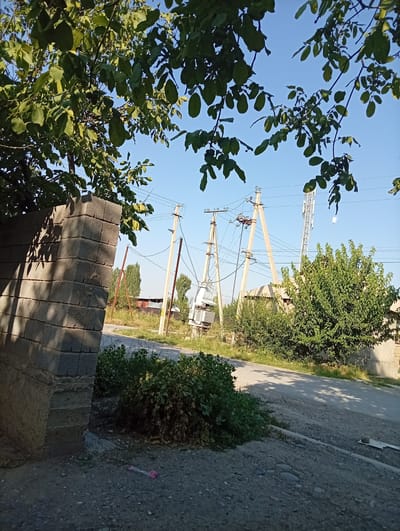 The image depicts a street scene in Kyzyl-Senir, Kyrgyzstan, on a clear day. In the foreground on the left, a grey brick pillar is visible, casting a shadow. A paved road with a gravel shoulder runs through the middle ground. To the right of the road, several utility poles with electrical wires are prominent, along with a large electrical transformer. A cellular tower stands further in the background, behind a residential structure. Lush green trees and bushes are scattered throughout the scene, with foliage from a tree overhead partially obscuring the sky. The ground appears to be a mixture of asphalt and gravel. A pink plastic object is lying on the gravel shoulder in the lower portion of the image.