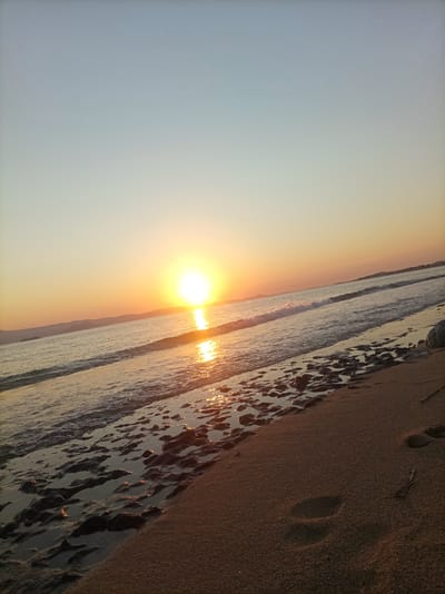 The image depicts a tranquil beach scene at sunset. The sun, a bright, luminous orb, is positioned low on the horizon, casting a warm, golden glow across the sky and the rippling water. Gentle waves are lapping at the sandy shore, leaving behind a wet, reflective surface dotted with small rocks and dark patches. In the foreground, footprints are visible in the sand, suggesting recent human presence. The background features a distant coastline with hills, silhouetted against the soft hues of the setting sun. The sky transitions from a pale blue at the top to a warm orange and yellow near the horizon. The overall atmosphere is peaceful and serene, capturing the beauty of a seaside sunset. The location is specified as Saint Arsenius, Greece.