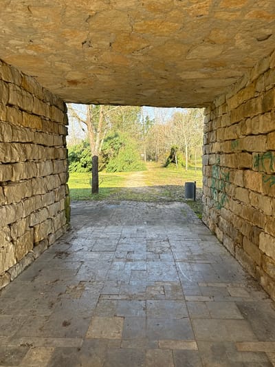 An outdoor stone passage in Mealhada, Portugal, opens into a park-like environment under clear daylight. The passage features walls and a ceiling constructed from irregularly shaped, light brown and tan stone blocks. The floor is paved with gray, unevenly laid stone tiles exhibiting wear and minor debris. Green graffiti marks are visible on the lower right interior wall of the passage. Beyond the opening, a grassy area extends, traversed by a narrow dirt path receding into the distance. A dark, cylindrical waste bin is positioned to the right of the passage exit. Various trees are present, including evergreens with dense foliage and deciduous trees with bare branches. No individuals are visible.