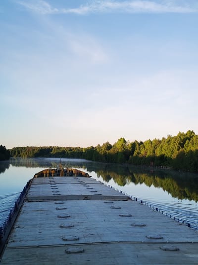 An expansive, serene view from the stern of a large, flat-decked cargo vessel, likely a barge, as it moves along a calm river or canal near Srednii Rubezh, Russia. The image depicts an early morning setting, with soft, golden sunlight illuminating the dense green forest lining both banks of the waterway. A delicate mist hovers over parts of the water surface, especially in the distant sections of the river, contributing to a tranquil atmosphere.
The grey, flat deck of the vessel, fitted with numerous circular, sealed hatches, occupies the foreground and midground. Blue railings or stanchions are visible along the side of the vessel, with ripples in the water indicating its forward motion. At the very back of the vessel, near the horizon line, a structure with yellow machinery, possibly part of a crane or steering mechanism, is discernible. The lush green trees, some brightly lit by the low sun, are clearly reflected in the still water. The sky above is a clear, light blue with a few faint, wispy clouds. No people are visible, and there is no discernible text in the image. The overall impression is one of peaceful navigation through a pristine natural environment.
