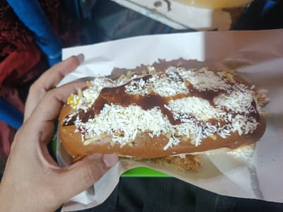 A hand holds a loaded hot dog on parchment paper. The bun is filled with a sausage, and topped with shredded cheese, chopped peanuts, and drizzled with a dark sauce. Additional toppings like ketchup and mustard are visible. A green dish holds the hot dog. The background includes clothing, a blue plastic chair, and a white surface.