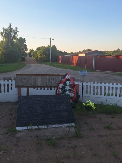 The image depicts a memorial located in the vicinity of Doktara Vitski Sel'ski Savet, Belarus. In the foreground, a black granite monument lists names, likely of fallen soldiers. A wreath with red and white flowers rests on top of the monument. Behind the monument, a decorative white fence encloses the memorial area. Further back, a large concrete structure displays the numbers "1941" and "1945" in large, weathered block letters. Beyond the memorial, a dirt road leads into the distance, flanked by lush green trees and grass. A tall, thin lamppost stands on the right side of the road. In the background, a red metal fence runs along the property line, and buildings with red roofs are visible. The sky is a clear, pale blue, suggesting it is either early morning or late afternoon. The overall scene is one of quiet commemoration and remembrance in a rural setting.