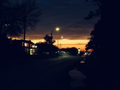 The image captures a twilight scene on a road in Salsk, Russia. The sky is a deep indigo at the top, transitioning to a warm orange and yellow band near the horizon, indicating sunset or sunrise. Silhouetted against the sky are the bare branches of trees on the left and a darker, denser tree on the right. A street lamp with a bright light illuminates the road in the mid-ground, and another fainter street lamp is visible further down. To the left, a building with lit windows is visible, suggesting it is a commercial establishment. The road surface is dark and reflects some of the ambient light, with a few puddles of water adding to the reflections. The overall atmosphere is dim and quiet, characteristic of a rural or suburban road at dusk. There are no people or animals visible in the frame.