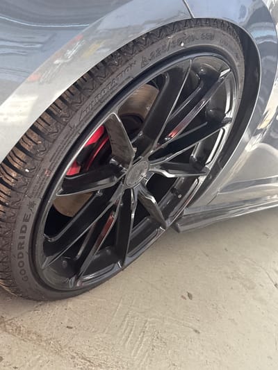 The image shows a close-up of a black alloy wheel with a red brake caliper visible behind it. The tire is a Goodride 225/35R19 88W. The car appears to be gray. The wheel is parked on a concrete surface, likely a road or parking lot, in Schkopau, Germany. The scene is outdoors, and the lighting suggests it is daytime. No people are visible in the image.