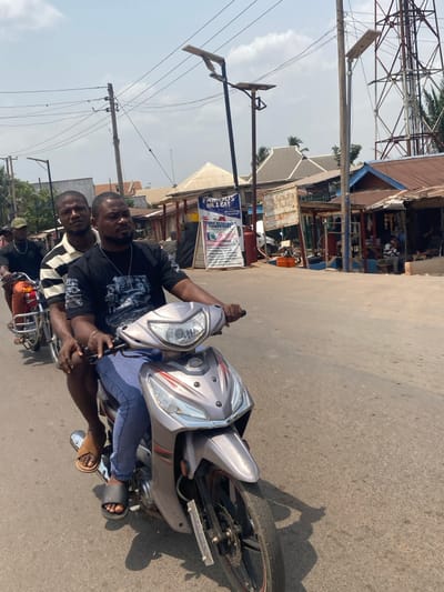 A daytime street scene in Afikpo, Nigeria, captures two adult males on a light gray scooter-style motorcycle, in motion on a paved road. The primary subject, driving, is a male wearing a dark t-shirt with a graphic design, blue jeans, and black slides. He holds the handlebars. A male passenger, wearing a horizontal striped t-shirt and light-colored sandals, rides directly behind the driver. Behind them, a third male operates a red motorcycle.
The environment includes overhead utility lines and multiple dark-colored poles supporting solar-powered streetlights. To the right, a series of single-story commercial buildings with corrugated metal roofs line the road. Prominent among these is a business identified by a large sign displaying "FAMOUS OIL & GAS" in blue text on a white background, positioned next to a red fuel dispenser. Another sign, "TARO RESOURCES COMPUTERS," is visible nearby. Orange jerry cans are situated on the ground near the "FAMOUS OIL & GAS" establishment. In the far background to the right, a tall metal lattice tower (likely a telecommunications mast) stands against a bright, hazy sky. Other individuals are present near the commercial structures. Strong shadows are cast on the road surface, indicating direct sunlight.