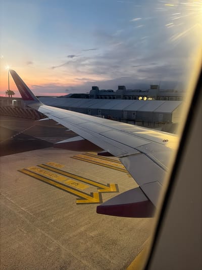 The image depicts an airplane wing with the Wizz Air logo visible on its tail fin, positioned on an airport tarmac. The scene captures a sunset with a sky painted in hues of blue, orange, and pink. Sunlight beams through the clouds, creating a lens flare effect in the upper left portion of the image.

In the background, an airport terminal building with multiple rows of air conditioning units is visible. Yellow arrows and markings are painted on the tarmac, indicating taxiway or parking guidance for aircraft. Text on these markings reads "E175" and other unreadable alphanumeric sequences. A palm tree is silhouetted against the colorful sunset in the lower right corner, suggesting a warm climate.

The time of day is sunset, characterized by the warm, golden light and colorful sky. The weather appears clear with scattered clouds. The overall impression is one of departure or arrival at an airport, with the airplane ready for its next flight. The image evokes a sense of travel and the beauty of a sunset over an airport.