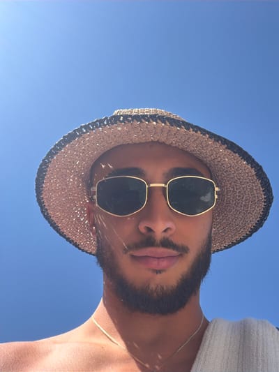 This media file is a low-angle, close-up photograph of a person wearing a straw hat and sunglasses, looking upwards against a clear blue sky. The subject has a beard and appears to be outdoors. The sun is bright, casting shadows from the hat onto the face, suggesting it is daytime. The person is wearing a white garment, possibly a shirt or sweater, and a thin gold chain necklace. The image has a relaxed and casual feel, potentially taken on a sunny day at a beach or other outdoor recreational area. The location context provided is Elaties Municipal Unit, Greece. There is no visible text in the image.