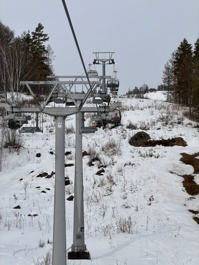 The scene depicts a ski slope in Novoabzakovo, Russia, under an overcast sky. A multi-column grey metal chairlift pylon, marked "3A," stands prominently in the foreground. Overhead, a steel cable supports several grey chairlifts. One chairlift in the mid-ground carries an individual clad in a pink jacket and blue pants, with a snowboard attached to their feet, ascending the slope. Other empty chairlifts are visible both behind and further up the line. The ground is partially covered in snow, revealing patches of dry, brown vegetation and scattered dark rocks. Visible ski tracks traverse the snowy surface. To the left, a cluster of bare deciduous trees stands against a backdrop of darker coniferous trees. To the right, the slope continues upwards, bordered by more coniferous trees along the horizon. Patches of exposed brown earth or mud are visible on the far right side of the image.