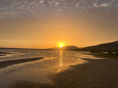 An eye-level, wide shot captures a serene sunset over a calm body of water, likely a lagoon or estuary, near Tarifa, Spain. The sun, a bright orange orb, is low on the horizon, casting a warm glow across the sky and reflecting vibrantly on the rippling water. The sky transitions from a soft orange near the sun to a pale yellow and then to a muted blue-grey with scattered clouds at the top of the frame. In the background, silhouetted hills form a gentle curve against the horizon. The foreground consists of a sandy beach with subtle wave patterns etched into the wet sand near the waterline, indicating a receding tide. There are two wooden posts on the right side of the frame, one with a sign attached, suggesting a designated area or marker. No people are visible in the image. The overall scene evokes tranquility and the natural beauty of a coastal sunset.