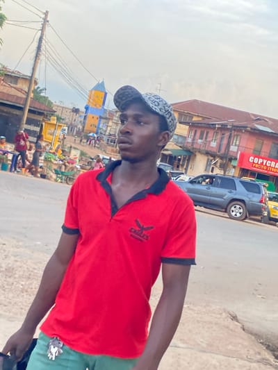 A man in a red shirt and patterned baseball cap stands on a street in Abofifia, Nigeria. Buildings and parked cars line the street. A sign for "COPYCRAFT PHOTO STUDIO" is visible on one of the buildings. A utility pole with wires stretches into the scene. People and market stalls populate the background. The sky is overcast.