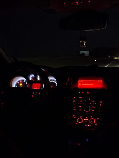 The image depicts the interior of a vehicle at night. The dashboard, instrument panel, and rearview mirror are visible, illuminated by red lights. The central display indicates "AUX Input Device" and a temperature of 21°C. The instrument panel includes the speedometer and other gauges, partially obscured by the steering wheel. The setting in Brestovets, Bulgaria is not discernible from the image. There are no people visible.