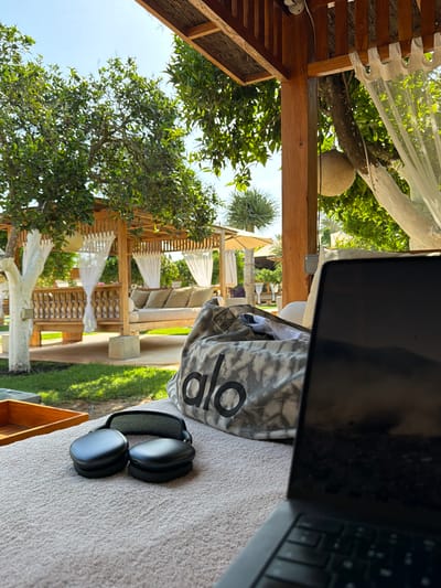 In the foreground, a laptop sits closed on a textured surface, its screen reflecting a dark, abstract image. To the left of the laptop, a pair of black headphones rests, their earcups facing upwards. Behind the headphones, a patterned duffel bag with the word "alo" printed in large black letters is visible. The background depicts an outdoor seating area with a wooden pergola providing shade. White curtains hang from the pergola, framing cushioned daybeds and seating arrangements. Lush green trees and a well-maintained lawn surround the area, with a bright blue sky visible overhead. The scene suggests a relaxed, outdoor workspace or lounging area, possibly in a resort or private residence. The specific location in Sant Joan de Labritja, Spain, is inferred from the context. There are no people visible in the image.