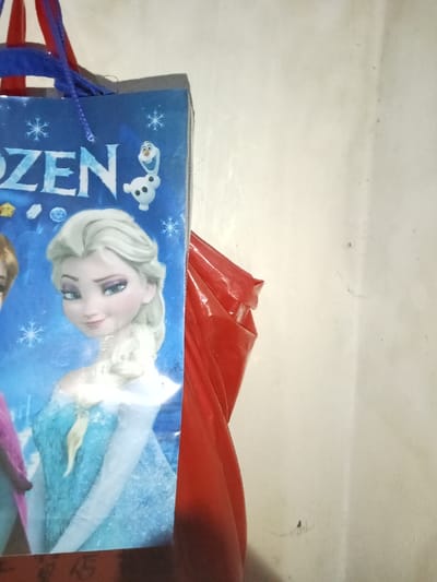 An indoor scene in Inxuba Yethemba Nu, South Africa, displays a blue paper gift bag positioned on the left side of the frame. The bag features characters from Disney's "Frozen," with Princess Elsa prominently centered, smiling, and wearing a light blue dress. Part of another character (likely Anna) is visible on Elsa's left. Above Elsa, the character Olaf is depicted. The word "ZEN" from "FROZEN" is visible at the top left, amidst snowflake patterns. Blue string handles are attached to the top of the bag. A red and purple strap is visible above the blue handles. To the right of the blue bag, a crumpled, glossy red plastic object is partially visible. The background consists of a plain, light-colored wall with minor marks. No human figures are present.
