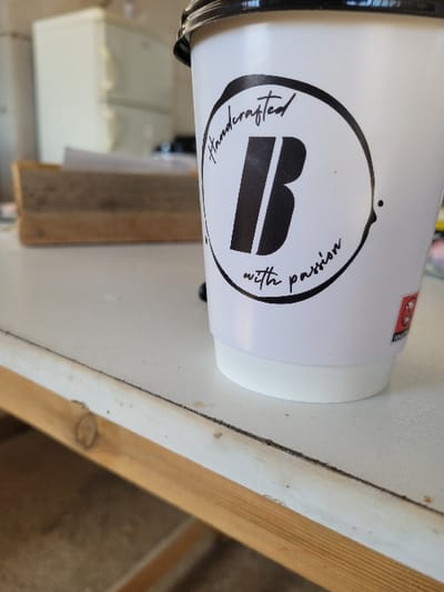 A white disposable coffee cup with a black lid is positioned on a light-colored, possibly white, surface. The cup features a black circular logo containing a bold black letter "B," with the script text "Handcrafted" above and "with passion" below. A small red graphic is partially visible on the right side of the cup. The surface exhibits minor wear and a faint dark line running horizontally. A wooden beam or structural element is visible beneath the surface. In the blurred background, a light-colored appliance, resembling a refrigerator or filing cabinet, is present, along with a wooden tray containing papers. The scene is an indoor environment in Ανδραβίδα, Greece. No people are visible, and no actions or interactions are occurring.