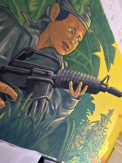 The mural depicts a close-up of a person holding a rifle, with a jungle-like environment surrounding them. The person has brown skin, dark hair, and is wearing a headscarf and a green uniform. Their gaze is directed downwards, and they appear to be in a defensive or alert posture. The rifle is held horizontally across the frame, with the person's hands gripping the weapon. Lush green foliage and yellow background fill the rest of the image. The mural is located on a wall in the historic center of Santa Marta, Colombia, specifically on the exterior of the former prison, La Modelo.